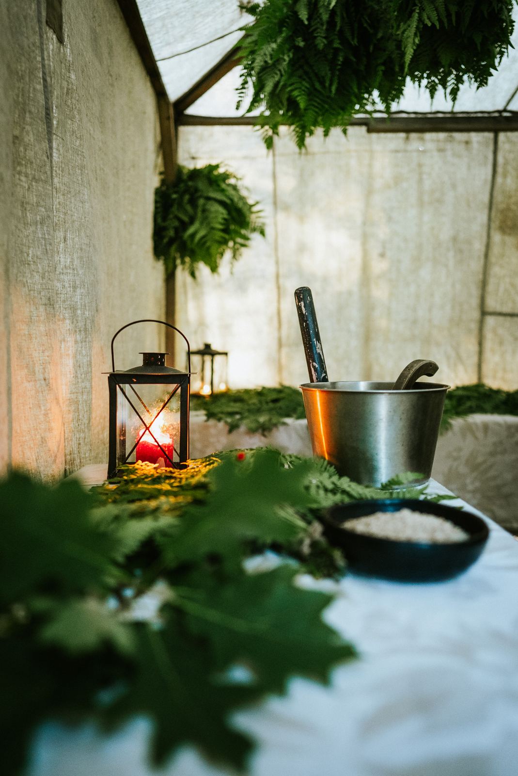 A candle lantern and a sauna bucket on a fern-laid table inside the tent