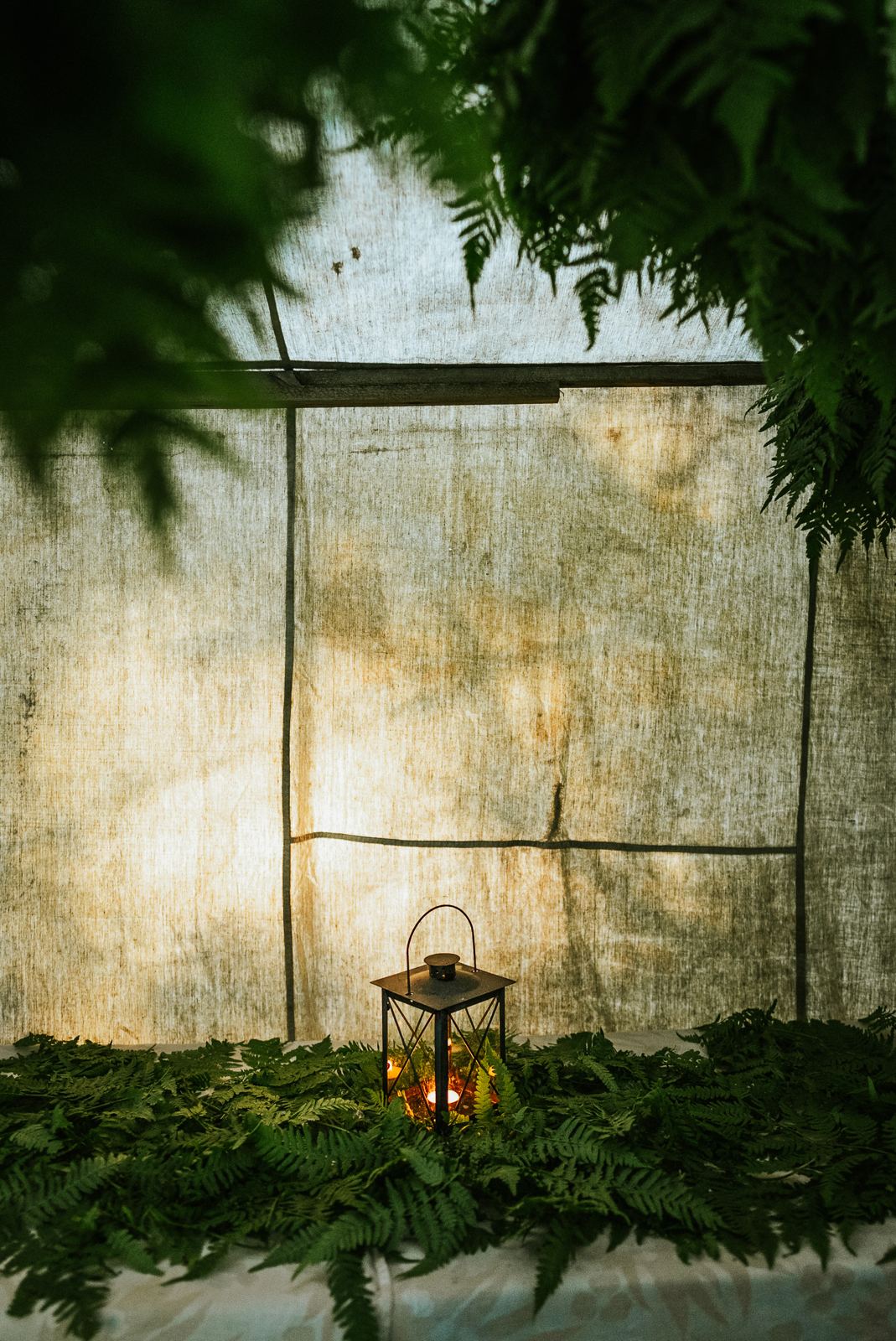 A candle lantern on a fern-covered table against a linen wall